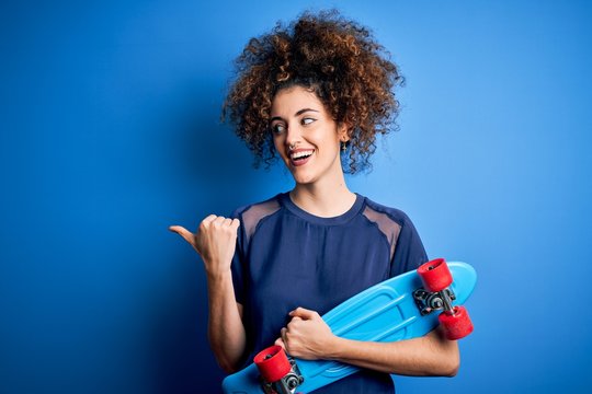 Young sporty woman with curly hair and piercing holding skate over blue background pointing and showing with thumb up to the side with happy face smiling