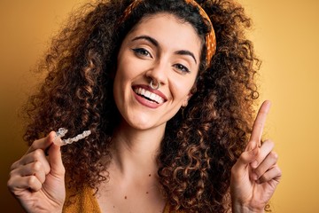 Young beautiful woman with curly hair and piercing holding dental aligner orthodontic surprised with an idea or question pointing finger with happy face, number one