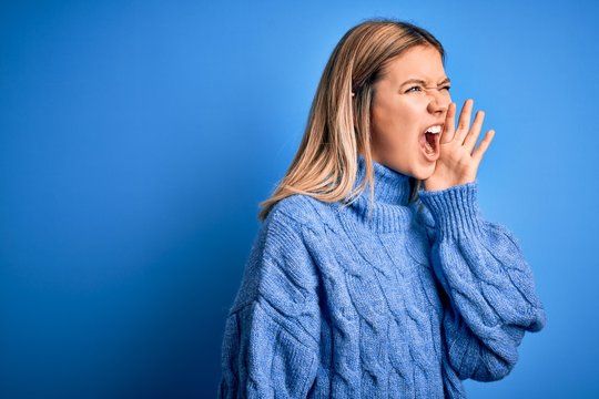 Young beautiful blonde woman wearing winter wool sweater over blue isolated background shouting and screaming loud to side with hand on mouth. Communication concept.