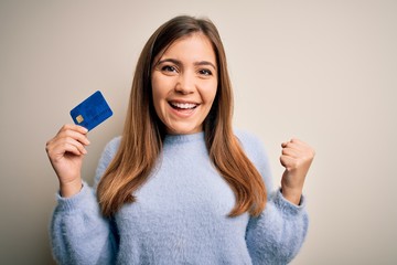 Young blonde woman holding credit card as payment over isolated background screaming proud and celebrating victory and success very excited, cheering emotion