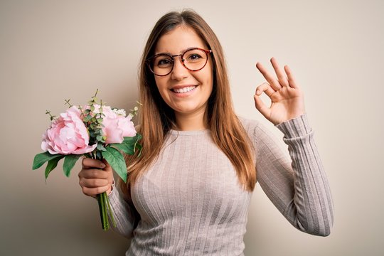 Young blonde woman holding beautiful romantic floral bouquet over isolated background doing ok sign with fingers, excellent symbol