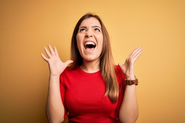 Beautiful young woman wearing casual red t-shirt over yellow isolated background celebrating mad and crazy for success with arms raised and closed eyes screaming excited. Winner concept