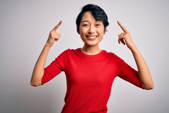 Young beautiful asian girl wearing casual red t-shirt standing over isolated white background smiling pointing to head with both hands finger, great idea or thought, good memory