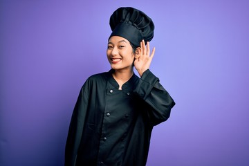 Young beautiful chinese chef woman wearing cooker uniform and hat over purple background smiling with hand over ear listening an hearing to rumor or gossip. Deafness concept.