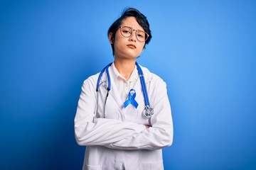 Young beautiful asian doctor girl wearing stethoscope and coat with blue cancer ribbon skeptic and nervous, disapproving expression on face with crossed arms. Negative person.