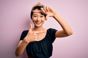 Fototapeta premium Young beautiful chinese woman wearing golden crown of king over isolated pink background smiling making frame with hands and fingers with happy face. Creativity and photography concept.