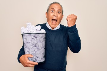 Senior grey-haired man holding full paper bin with crumpled papers over white background screaming...