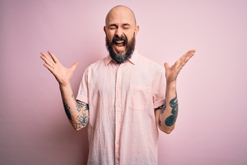 Fototapeta premium Handsome bald man with beard and tattoo wearing casual shirt over isolated pink background celebrating mad and crazy for success with arms raised and closed eyes screaming excited. Winner concept
