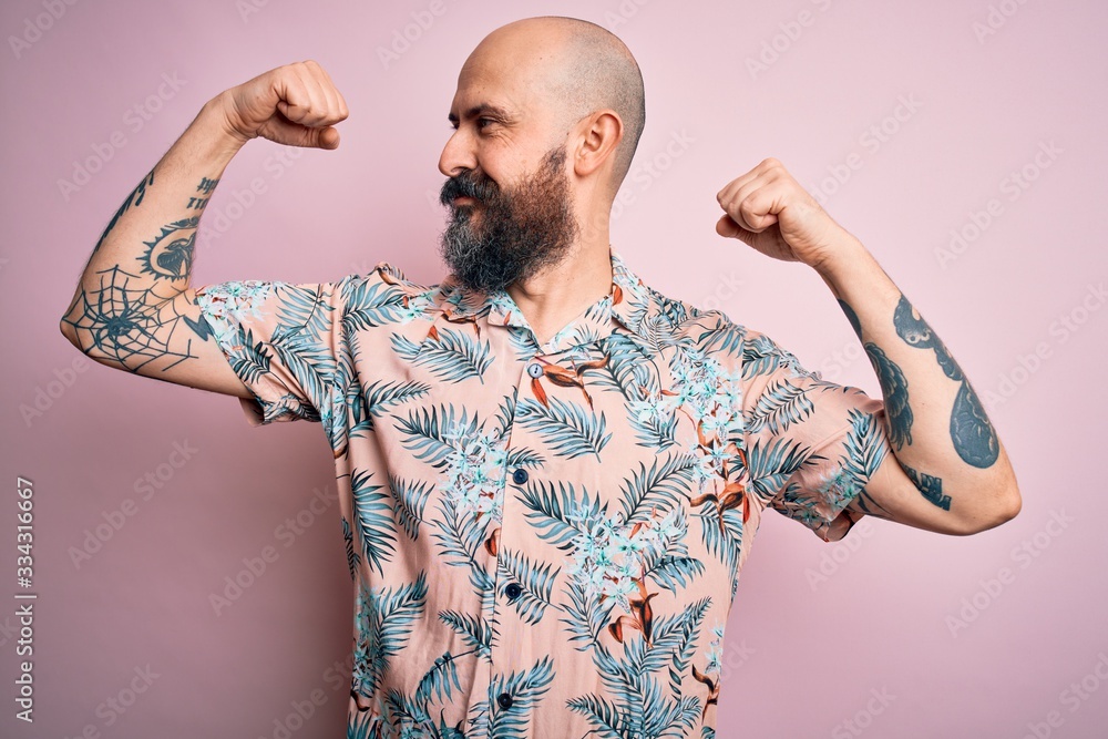 Sticker Handsome bald man with beard and tattoo wearing casual floral shirt over pink background showing arms muscles smiling proud. Fitness concept.