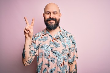 Naklejka premium Handsome bald man with beard and tattoo wearing casual floral shirt over pink background showing and pointing up with fingers number two while smiling confident and happy.