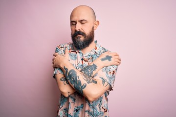 Handsome bald man with beard and tattoo wearing casual floral shirt over pink background Hugging oneself happy and positive, smiling confident. Self love and self care