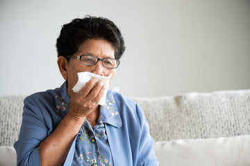 Sick asian old woman using tissue paper close mouth while cough, sitting on sofa at home. Senior healthcare concept.