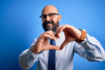 Handsome business bald man with beard wearing elegant tie and glasses over blue background smiling in love doing heart symbol shape with hands. Romantic concept.