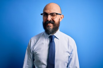 Handsome business bald man with beard wearing elegant tie and glasses over blue background winking...