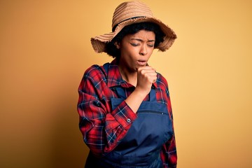 Young African American afro farmer woman with curly hair wearing apron and hat feeling unwell and coughing as symptom for cold or bronchitis. Health care concept.