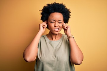 Young beautiful African American afro woman with curly hair wearing casual t-shirt covering ears with fingers with annoyed expression for the noise of loud music. Deaf concept.