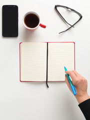 Female hand holding a blue pen over a blank notebook, with mobile, cup of coffee, glasses, white background Top view mockup smartphone black screen