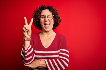 Middle age beautiful curly hair woman wearing casual striped sweater over red background smiling with happy face winking at the camera doing victory sign. Number two.