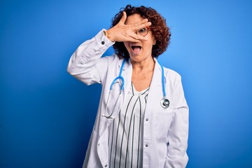 Middle age curly hair doctor woman wearing coat and stethoscope over blue background peeking in shock covering face and eyes with hand, looking through fingers with embarrassed expression.