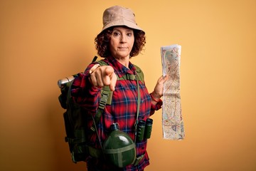 Fototapeta na wymiar Middle age curly hair hiker woman hiking wearing backpack and water canteen holding city map pointing with finger to the camera and to you, hand sign, positive and confident gesture from the front