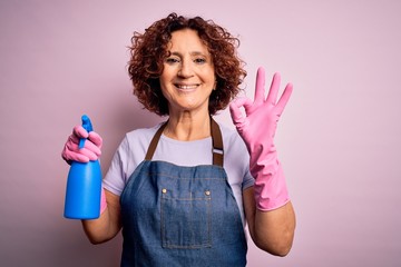 Middle age curly hair woman cleaning doing housework wearing apron and gloves using spayer doing ok sign with fingers, excellent symbol