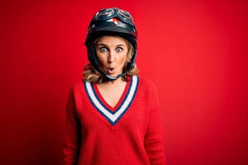 Middle age beautiful blonde motorcyclist woman wearing moto helmet over red background afraid and shocked with surprise expression, fear and excited face.