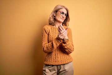 Middle age beautiful blonde woman wearing casual sweater and glasses over yellow background...
