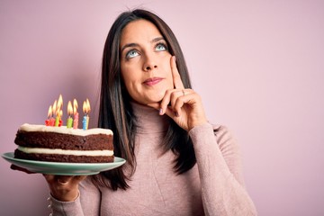 Young woman with blue eyes holding birthday cake with candles over pink background serious face...