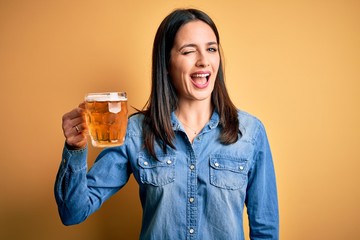 Young woman with blue eyes drinking jar of beer standing over isolated yellow background winking...