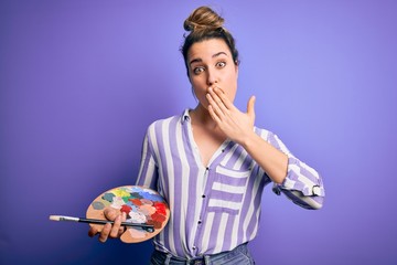 Fototapeta premium Young beautiful blonde artist woman painting using brush and palette over purple background cover mouth with hand shocked with shame for mistake, expression of fear, scared in silence, secret concept