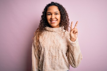 Young beautiful woman with curly hair wearing casual sweater standing over pink background showing and pointing up with fingers number two while smiling confident and happy.