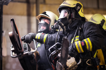 Group of professional firefighters wearing full equipment, oxygen masks, and emergency rescue tools, circular hydraulic and gas saw, axe, and sledge hammer. smoke and fire trucks in the background.