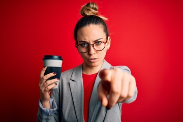 Young beautiful blonde businesswoman with blue eyes wearing glasses drinking cup of coffe pointing with finger to the camera and to you, hand sign, positive and confident gesture from the front