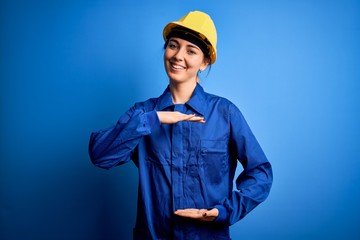 Young beautiful worker woman with blue eyes wearing security helmet and uniform gesturing with hands showing big and large size sign, measure symbol. Smiling looking at the camera. Measuring concept.