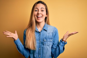 Young beautiful blonde woman with blue eyes wearing denim shirt over yellow background celebrating crazy and amazed for success with arms raised and open eyes screaming excited. Winner concept