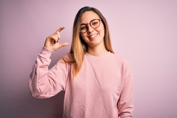 Beautiful blonde woman with blue eyes wearing sweater and glasses over pink background smiling and confident gesturing with hand doing small size sign with fingers looking and the camera. Measure