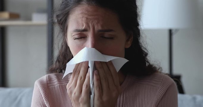 Ill sick young woman sneezing in tissue, blowing runny nose. Millennial lady having flu coronavirus cold symptoms at home. Covid 19 virus infection, flu and seasonal allergy concept. Close up view