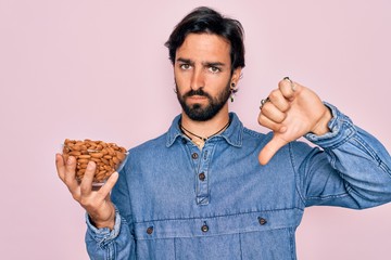 Young handsome hispanic man with bear holding natural ingredient healthy almonds with angry face,...