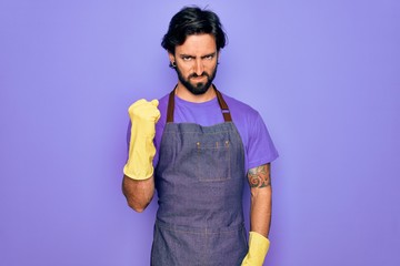 Young handsome hispanic clenaer man wearing housework apron and washing gloves angry and mad raising fist frustrated and furious while shouting with anger. Rage and aggressive concept.