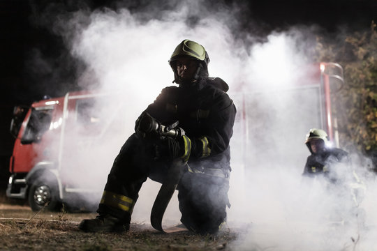 Team of professional firefighters holding water hose in front of a firetruck with smoke in the air. Half silhouette.