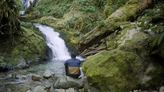Male sitting peacefully relaxed in front of beautiful waterfall in pacific northwest rain forest with crystal clear blue pool