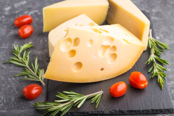 Various types of cheese with rosemary and tomatoes on black slate board on a black concrete background. Side view, close up.