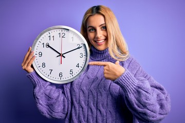 Young beautiful blonde woman doing countdown holding big clock over purple background very happy pointing with hand and finger