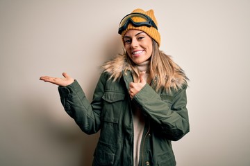 Young brunette skier woman wearing snow clothes and ski goggles over white background Showing palm hand and doing ok gesture with thumbs up, smiling happy and cheerful