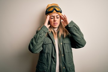 Young brunette skier woman wearing snow clothes and ski goggles over white background with hand on headache because stress. Suffering migraine.