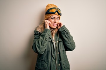 Young brunette skier woman wearing snow clothes and ski goggles over white background covering ears with fingers with annoyed expression for the noise of loud music. Deaf concept.