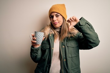 Young beautiful brunette woman wearing snow clothes drinking mug of coffee with angry face,...