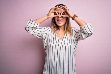 Young beautiful woman wearing casual striped t-shirt and glasses over pink background doing ok gesture like binoculars sticking tongue out, eyes looking through fingers. Crazy expression.