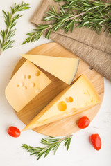 Various types of cheese with rosemary and tomatoes on wooden board on a white wooden background. Top view, close up.