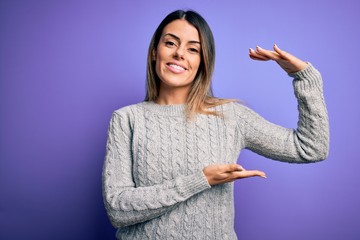 Young beautiful woman wearing casual sweater standing over isolated purple background gesturing with hands showing big and large size sign, measure symbol. Smiling looking at the camera. Measuring